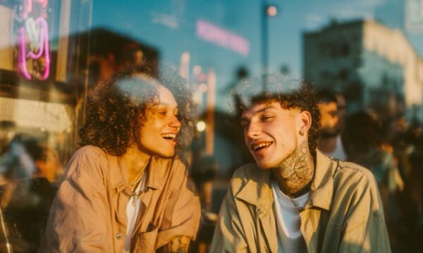 A candid photo of two people sitting outside a cocktail bar at dusk, one laughing with a glass in hand, the other leaning forward. Neon signs reflected in windows behind, realism in body language. Slight grain, cinematic color palette, unretouched and honest. --profile nflquj2 --v 7 Job ID: db6a9704-783a-493c-ba54-468448fb1f0f