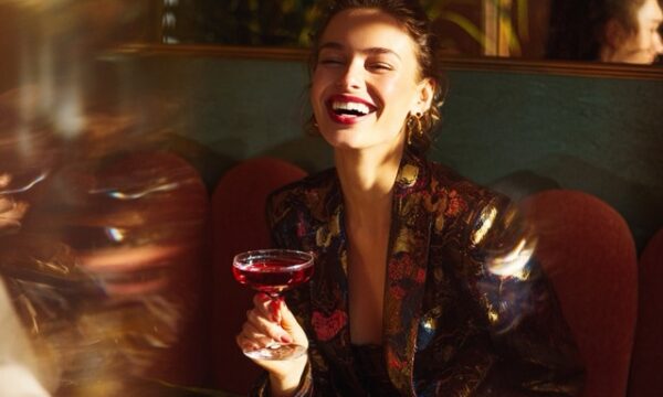 Fashion editorial photo of a stylish woman laughing with friends in a high-end cocktail bar. She wears a modern, classy, expensive fashionable streetwear outfit, captured in motion with painterly blur and ambient light streaks. Her hand holds a coupe glass with a deep red cocktail, glinting under golden reflections from glass and polished surfaces. The setting features rich terracotta and warm clay tones from the furniture and floor, contrasted with muted olive and forest green walls or plants. The lighting is bright but natural not clearly day or night soft glows, motion haze, and shallow depth of field. Inspired by Jacquemus campaign styling and elegant surrealism. Editorial mood, not nightlife energy. No logos. --v 7 Job ID: efa2eb1b-9969-4ec0-b885-b9614361f08e
